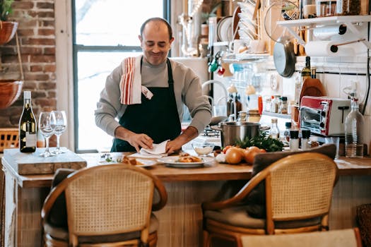 Positive middle aged male cooking food in bright room near counter with plates with pasta near onion and herbs near kitchenware and wine bottle near glasses and chairs near window
